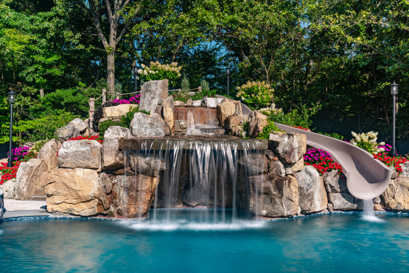 Nicosia A - Cream Ridge, NJ - A large rock waterfall with a slide in the middle of a swimming pool surrounded by lush greenery and colorful flowers