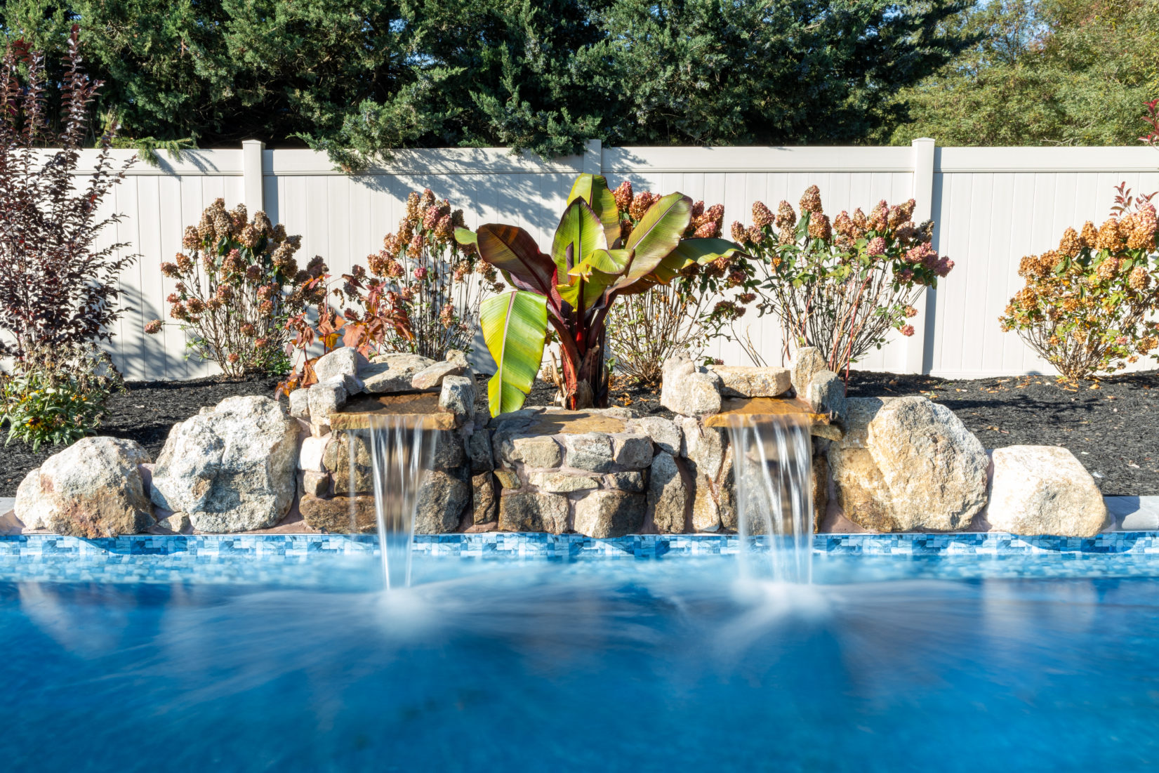 Genesis P -  - A swimming pool with a waterfall feature surrounded by plants and a white fence