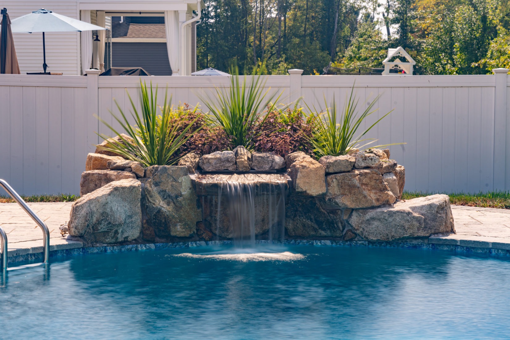 Trado A - Bel Aire, MD - A swimming pool with a waterfall in the middle of it surrounded by plants and a white fence