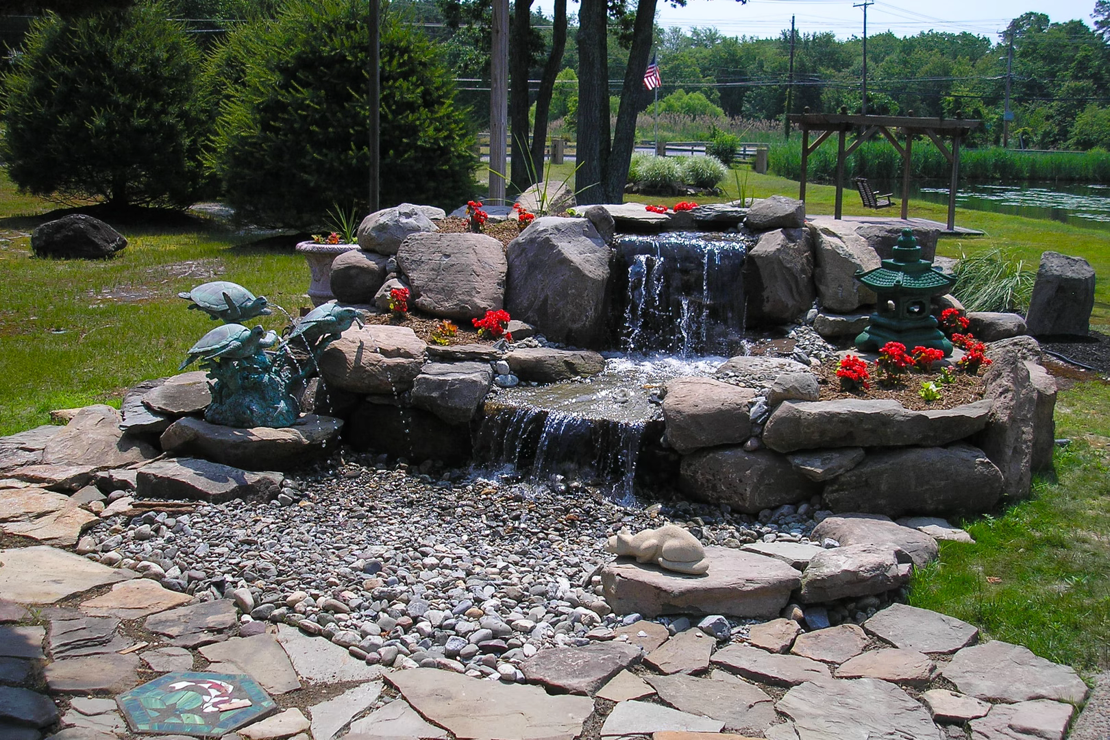 Two-tiered natural stone boulder waterfall cascading into a pebble basin near lush garden plants