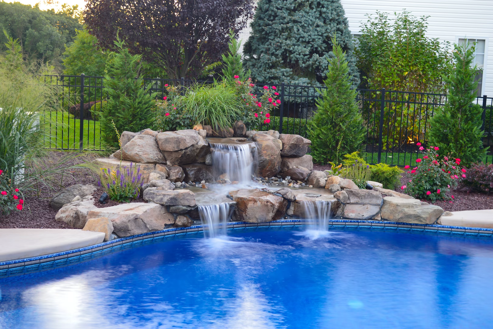 Multi-tiered natural stone boulder waterfall cascading into a pool surrounded by lush garden plants