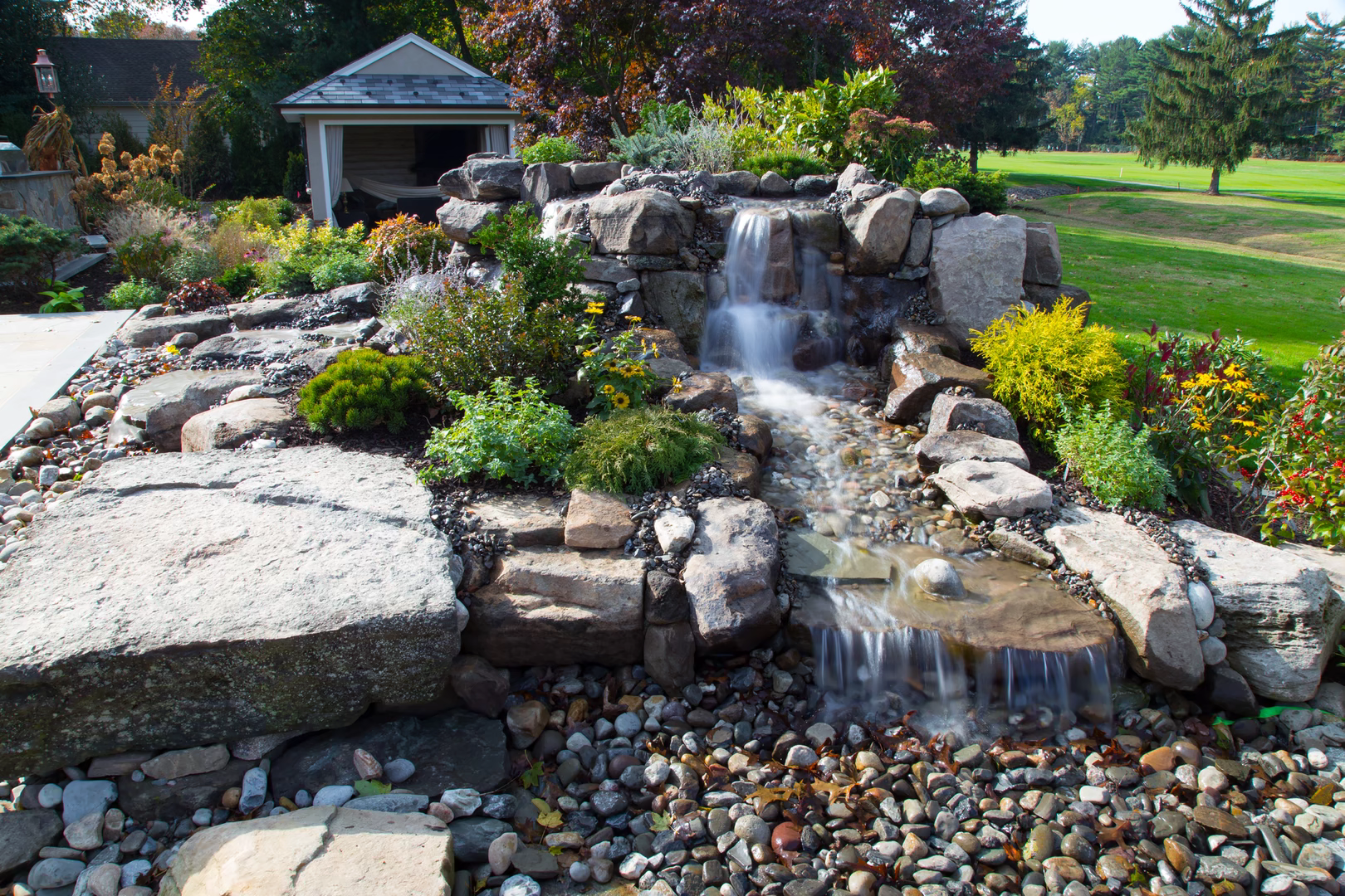 Two-tiered natural stone boulder waterfall cascading into a pebble basin with lush garden landscaping