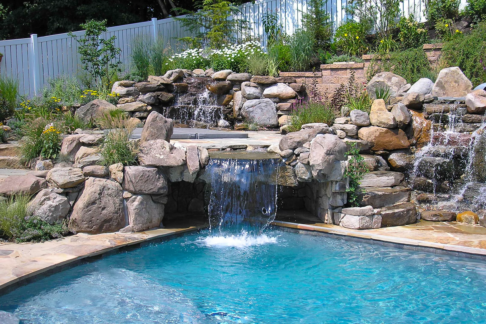 Multi-tiered natural stone boulder waterfalls cascading into a swimming pool with lush garden landscaping