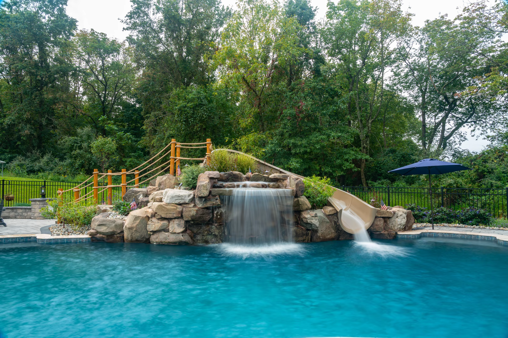 Two-tiered natural stone boulder waterfall cascading into a pool surrounded by lush green trees
