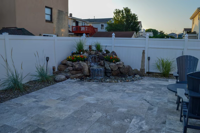 Atera B - Brigantine, NJ - A backyard with a stone patio and a waterfall feature surrounded by plants and flowers