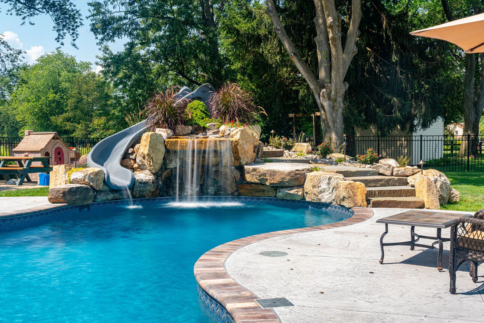 Multi-tiered natural stone boulder waterfall cascading into a pool with lush plants and slide