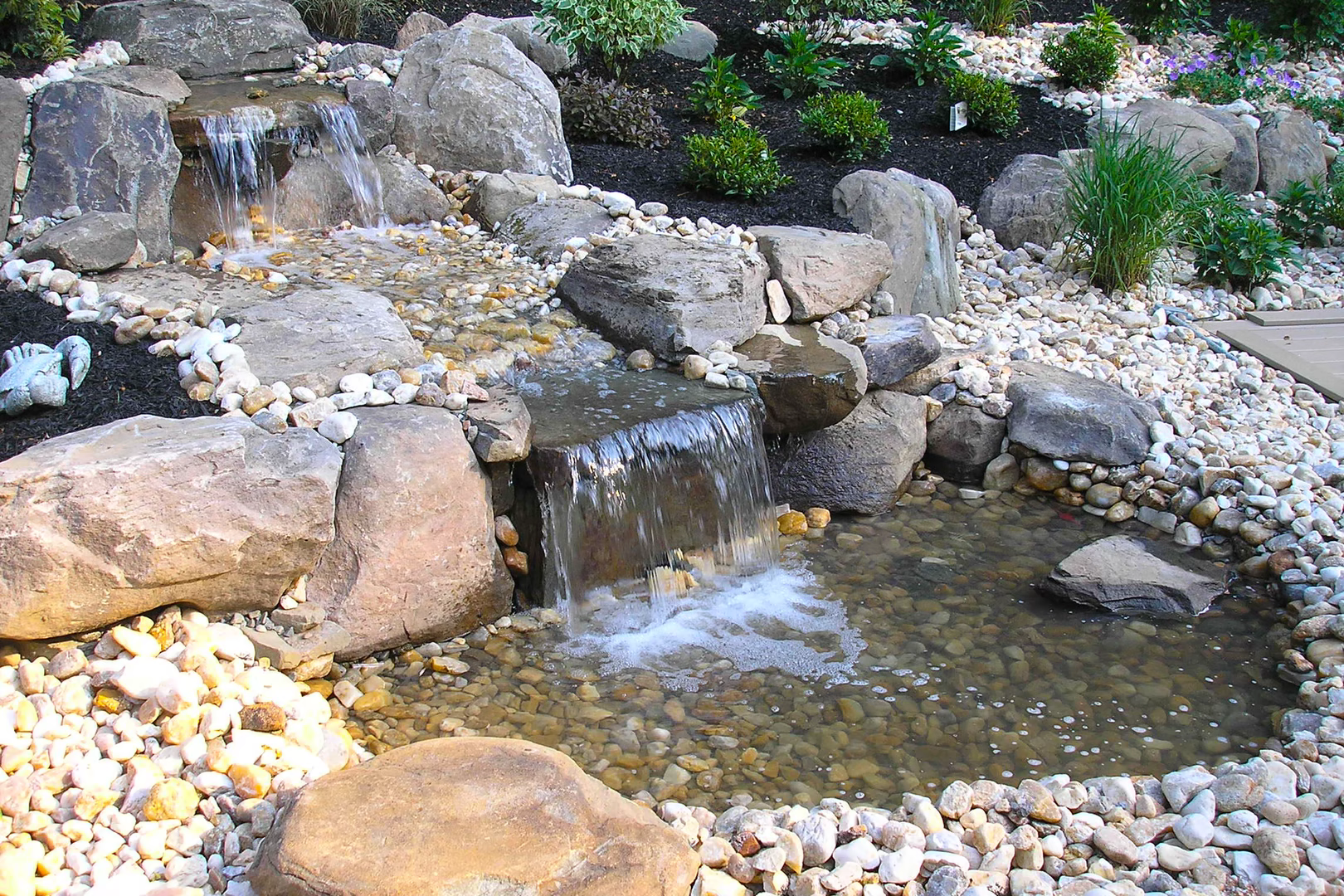 Two-tiered natural stone boulder waterfall cascading into a pebble pool surrounded by lush plants