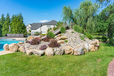 Bagley F - Williamstown, NJ - A backyard with a large rock wall and a slide, surrounded by lush green grass and trees
