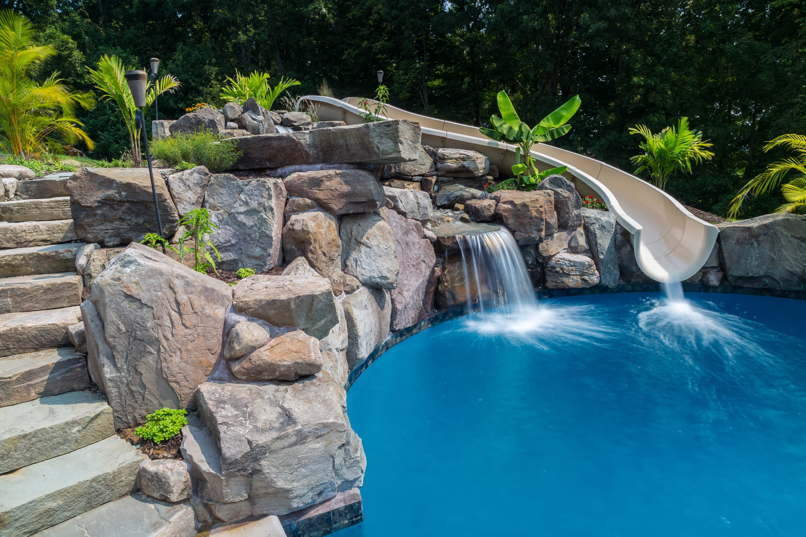 Multi-tiered natural stone boulder waterfall cascading into a pool surrounded by lush tropical landscaping