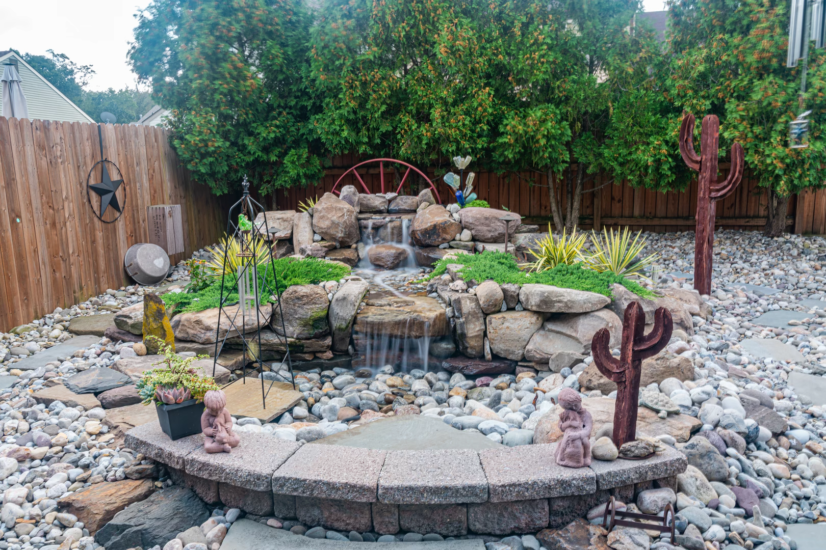 Multi-tiered natural stone boulder waterfall cascading into a pebble basin surrounded by lush green plants