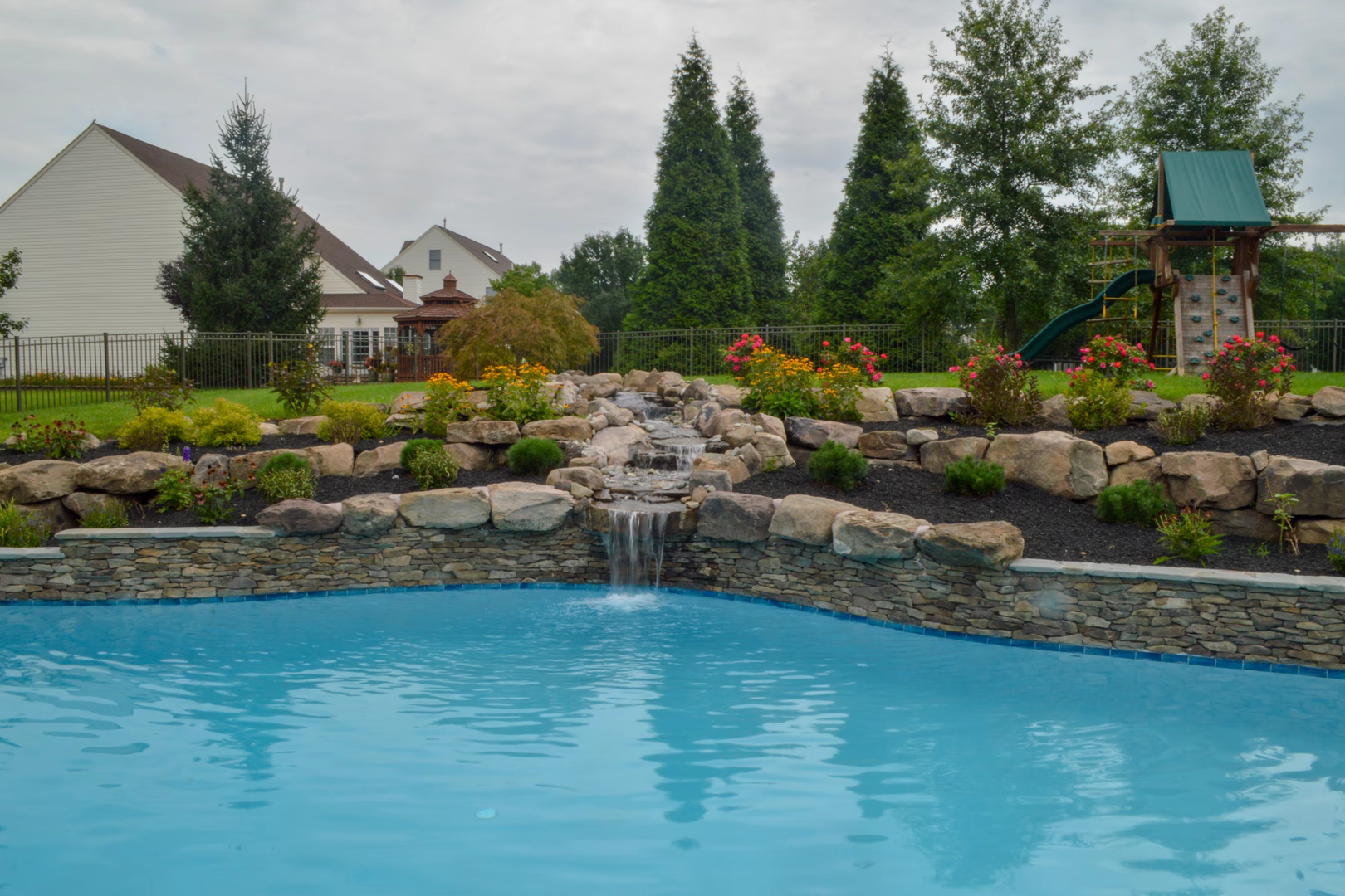 Multi-tiered natural stone boulder waterfall cascading into a pool surrounded by colorful garden plants