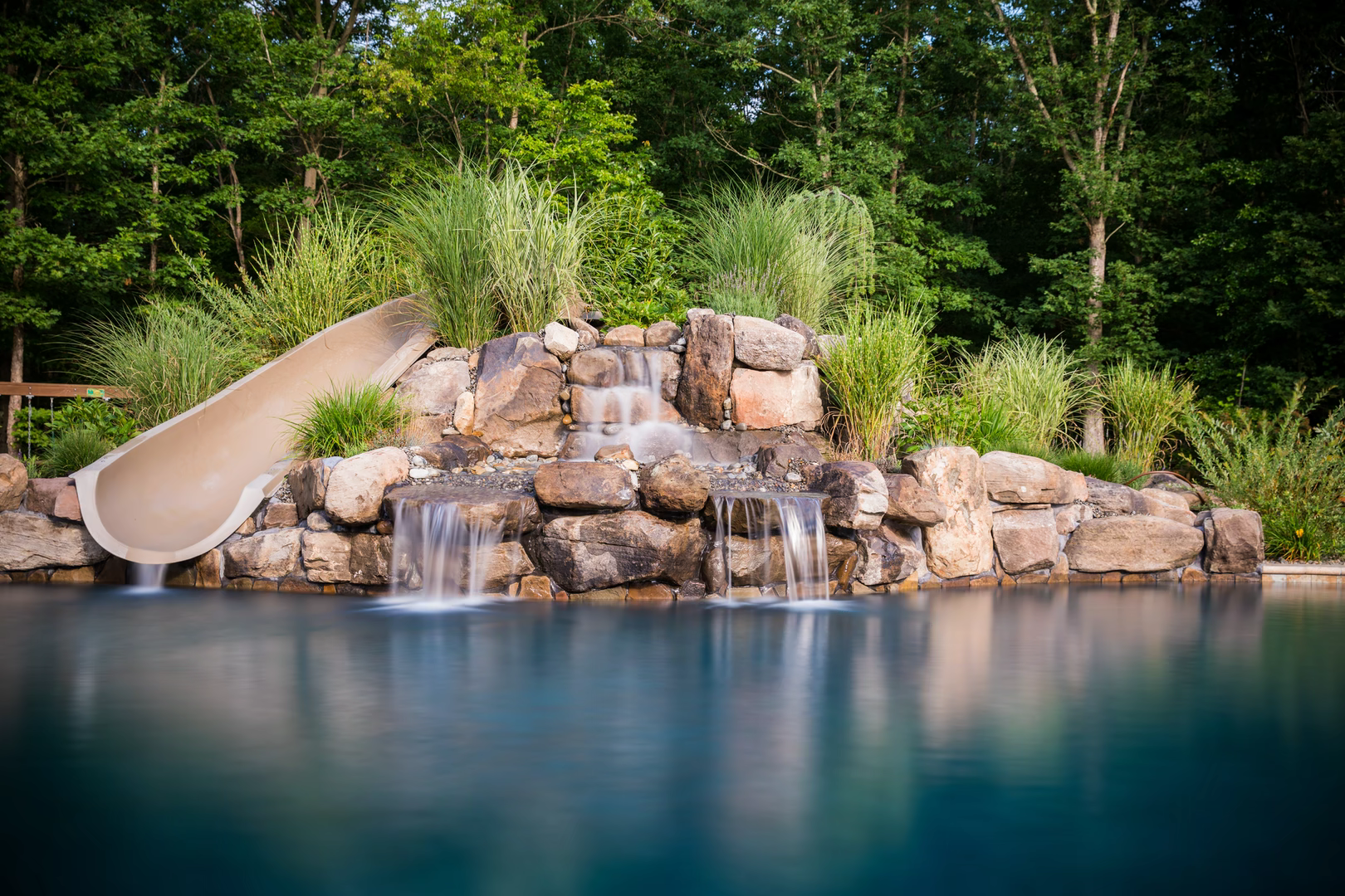Multi-tiered natural stone boulder waterfalls cascading into a swimming pool surrounded by lush greenery