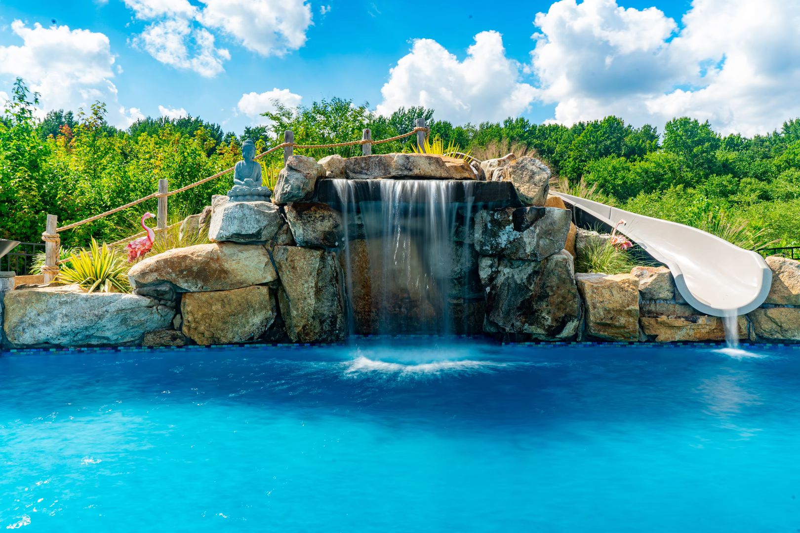 Single-tier boulder waterfall cascading into a blue pool with lush greenery and waterslide