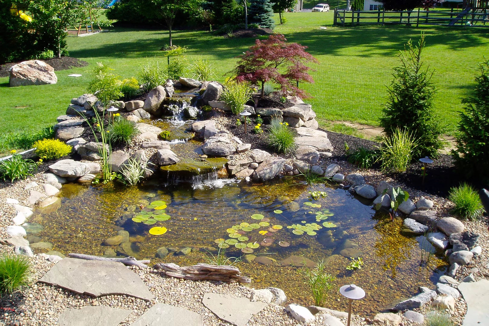Multi-tiered natural stone boulder waterfall cascading into a garden pond surrounded by lush plants
