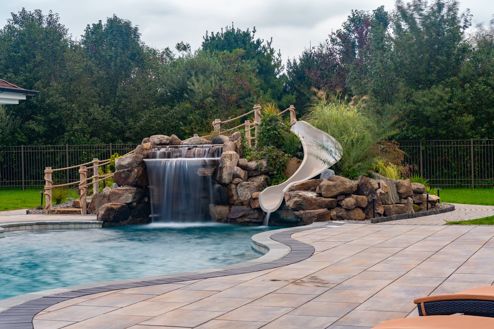 Double-tiered natural stone boulder waterfall cascading into a pool with lush landscaping and slide