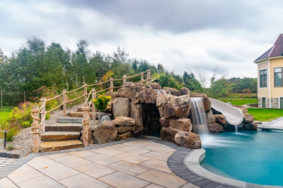 Bruar B - Millstone, NJ - A backyard with a pool and a waterfall feature, surrounded by lush greenery and a house in the background