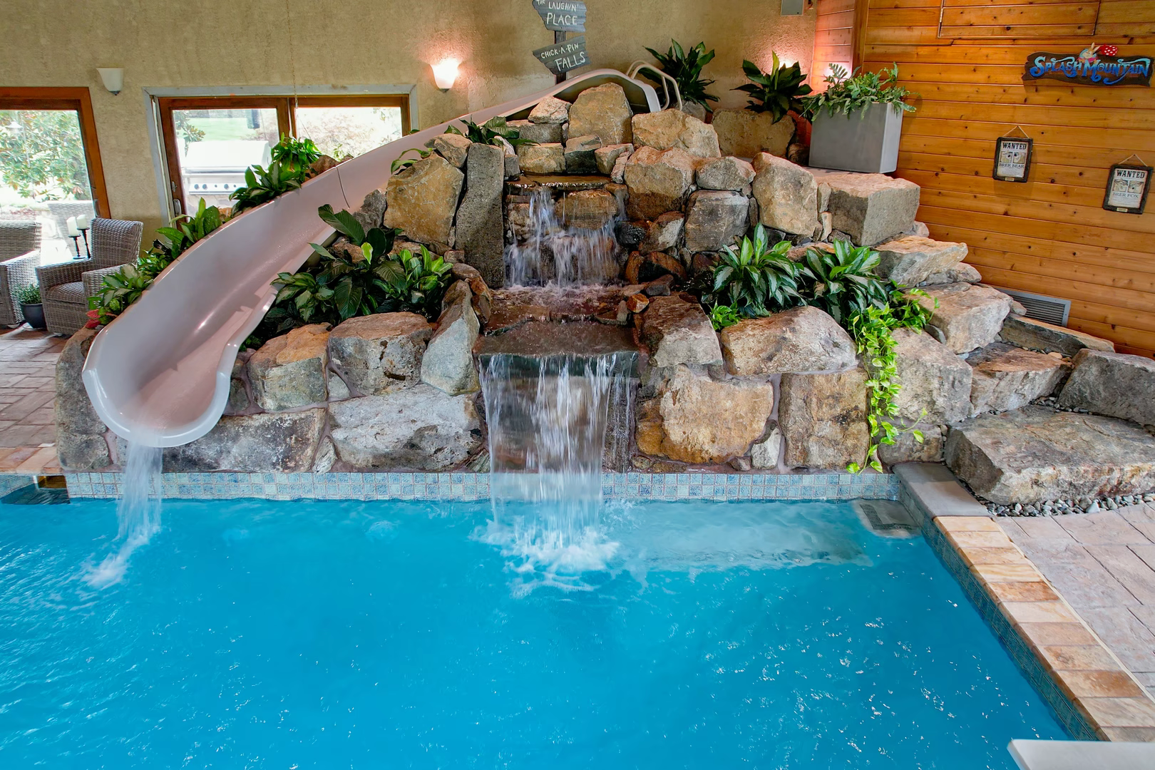 Two-tiered natural stone boulder waterfall cascades into a clear pool with lush indoor plants