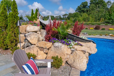 Chania E - Hamilton, NJ - A rock wall with a water feature next to a swimming pool, surrounded by lush greenery and colorful flowers