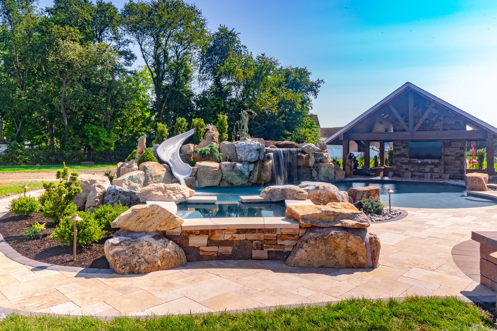 Multi-tiered natural stone boulder waterfall cascading into a pool with slide and lush landscaping