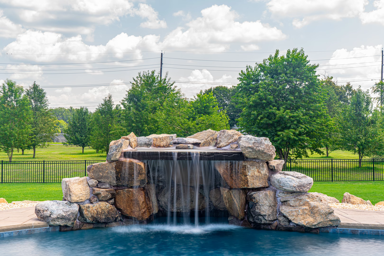 Single-tier natural stone boulder waterfall with curtain flow cascading into a pool near green trees
