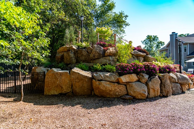 Copper M - Cream Ridge, NJ - A large rock retaining wall with flowers in the foreground and a house in the background