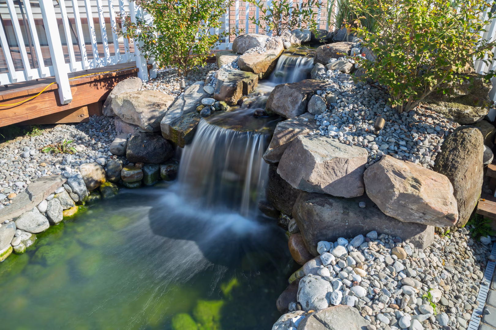 Two-tiered natural stone boulder waterfall cascading into a clear pool surrounded by garden plants