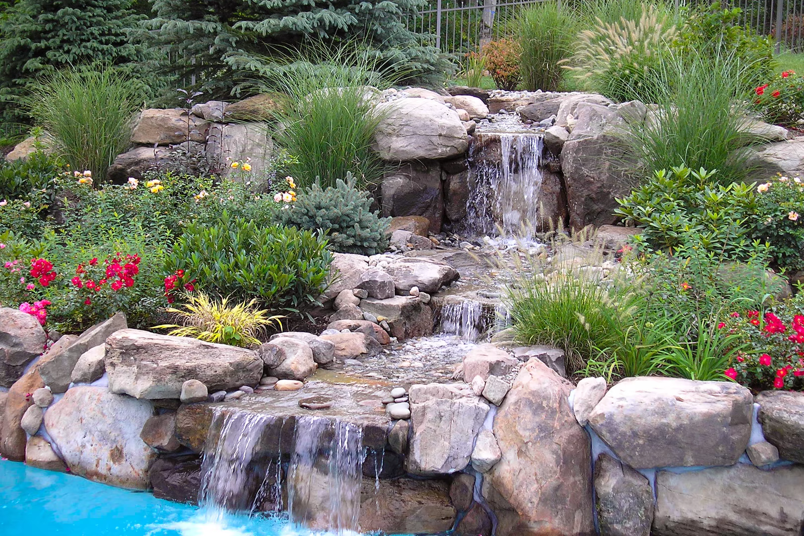 Three-tiered natural stone boulder waterfall cascading into a pool surrounded by lush garden plants