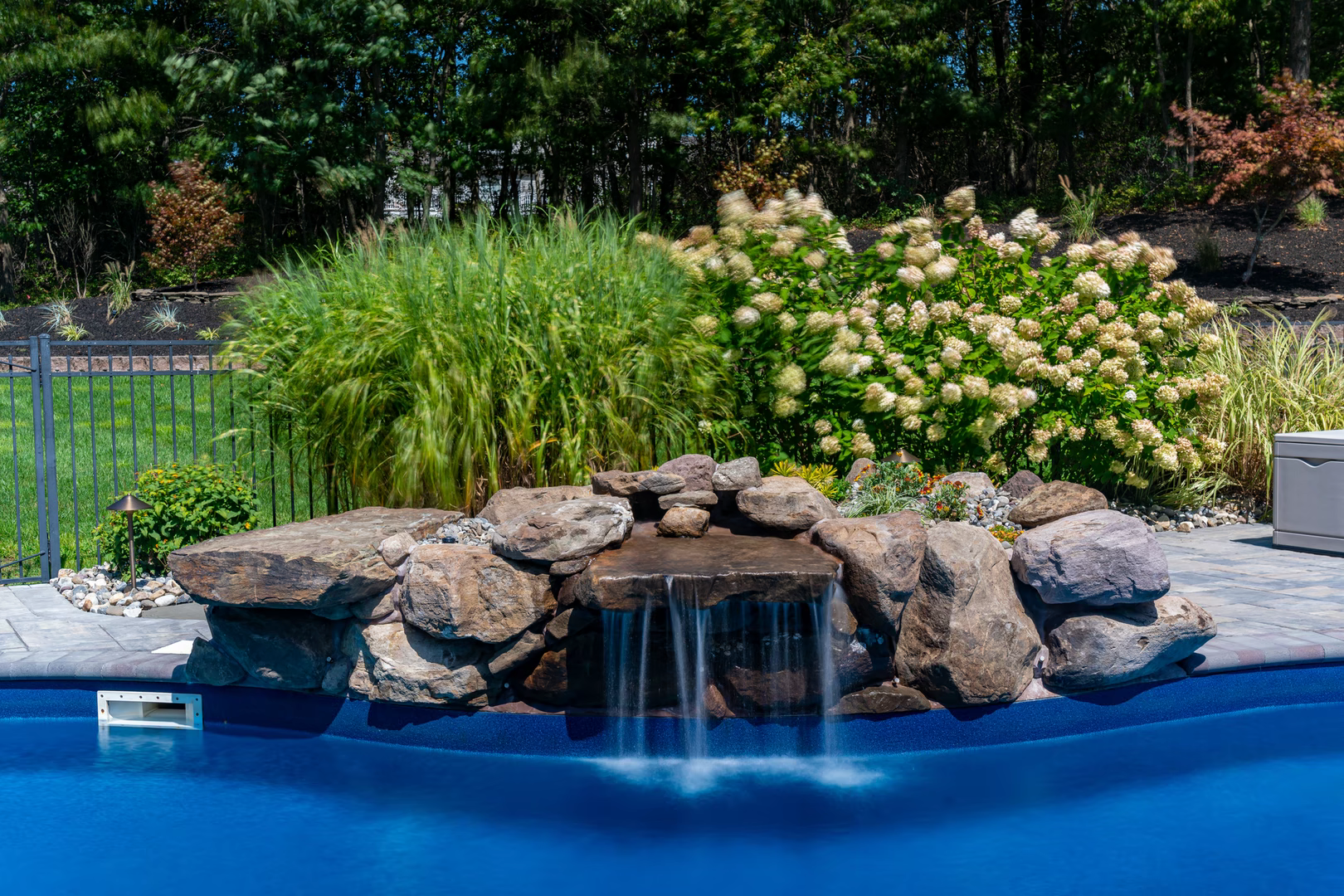 Two-tiered natural stone waterfall cascading into a blue pool surrounded by lush garden greenery