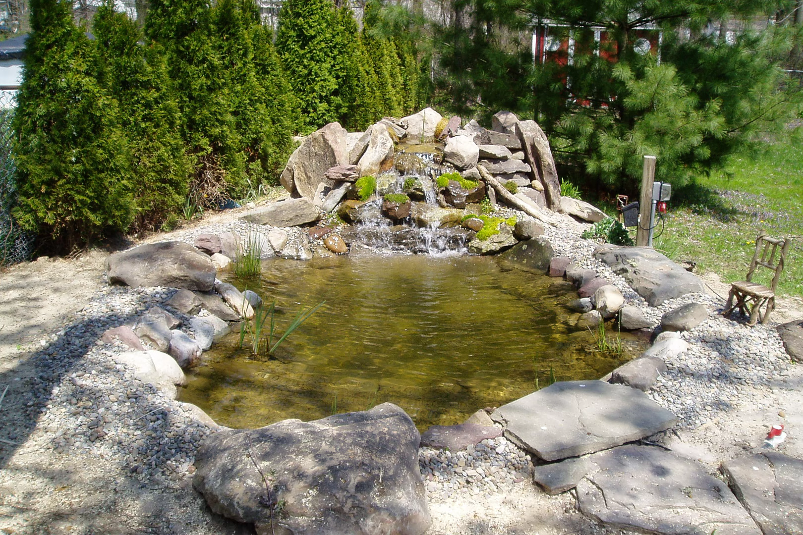 Natural stone boulder waterfall cascading into a garden pond surrounded by evergreens