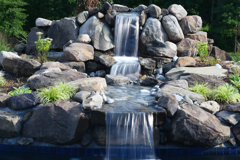Natural stone boulder waterfall cascading into a pool with lush garden plants