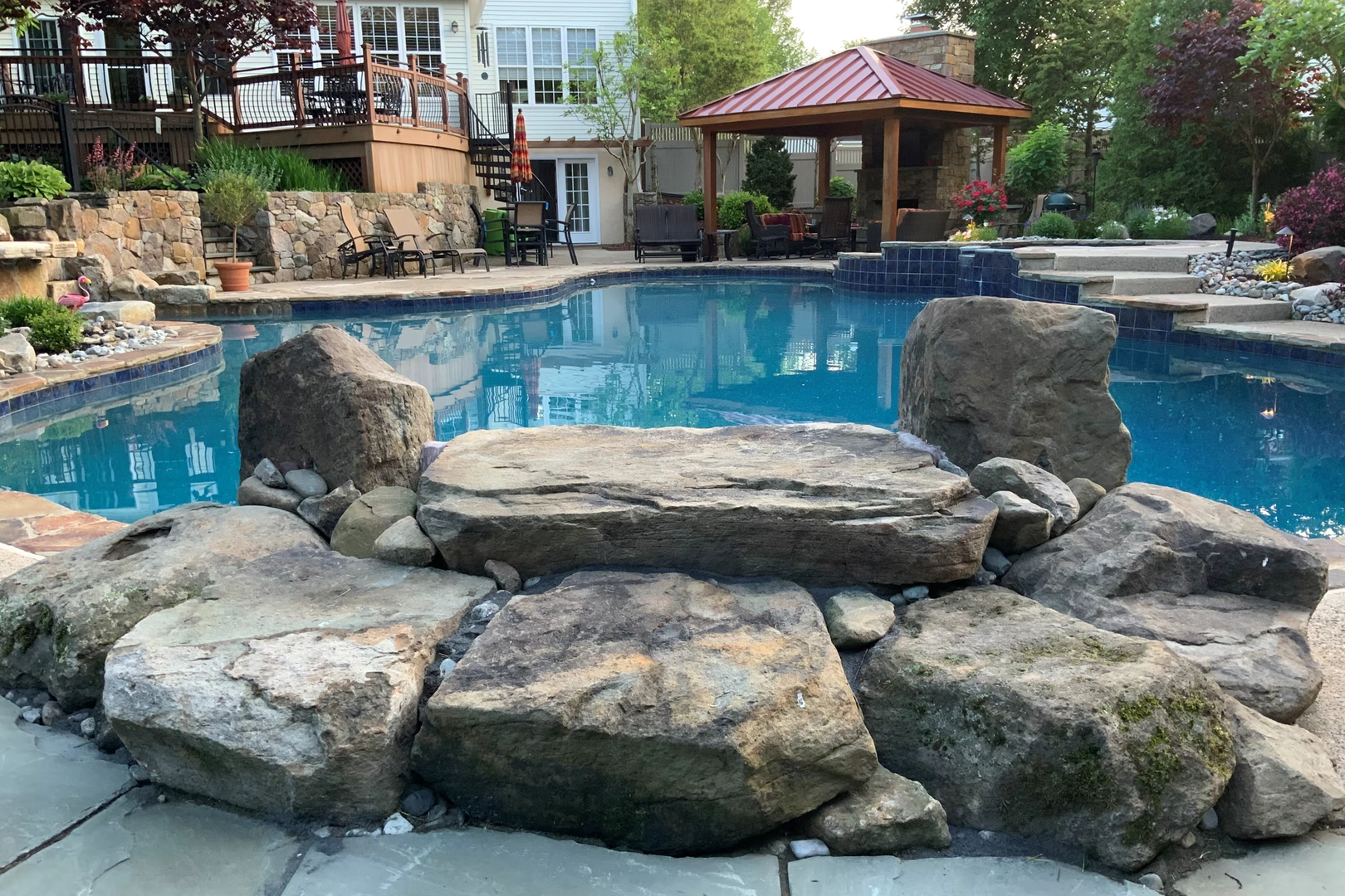 Multi-tiered natural stone boulder waterfall cascading into a backyard pool with lush garden plants
