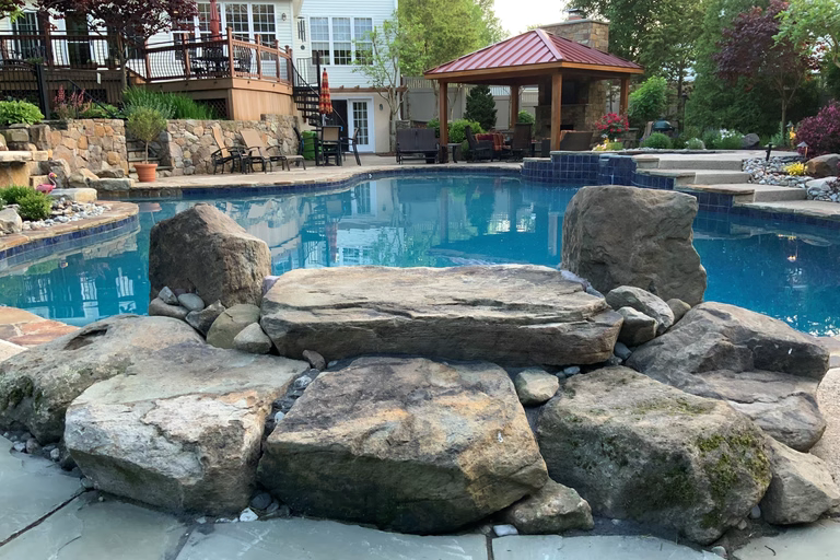 Natural stone boulder waterfall cascading into a backyard pool with lush garden plants