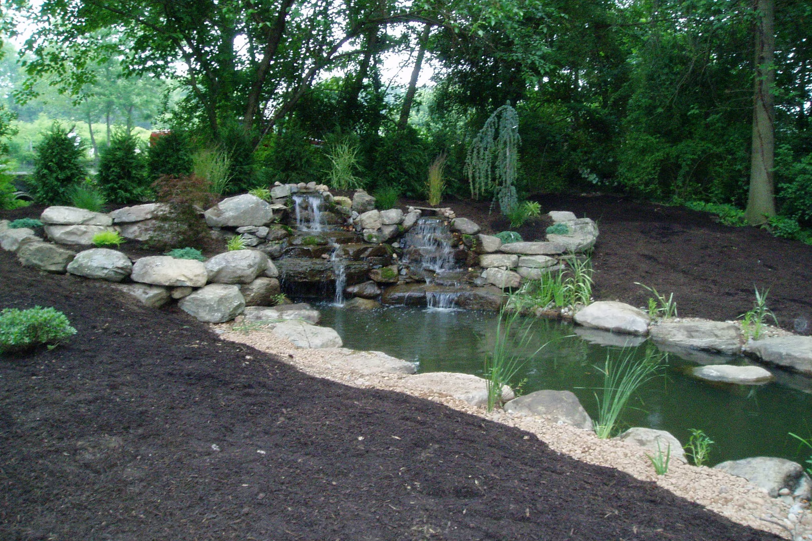 Natural stone boulder waterfall cascading into a pond surrounded by lush garden plants