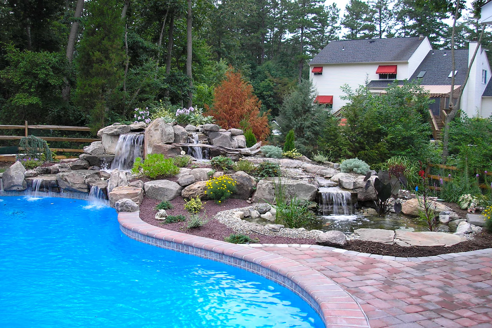 Multi-tiered natural stone boulder waterfalls spill into a freeform pool surrounded by lush greenery