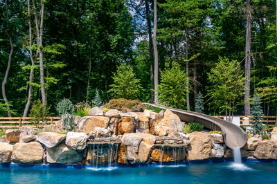 Gavarnie A - Monkton, MD - A large rock waterfall with a slide in the middle of a swimming pool surrounded by lush greenery