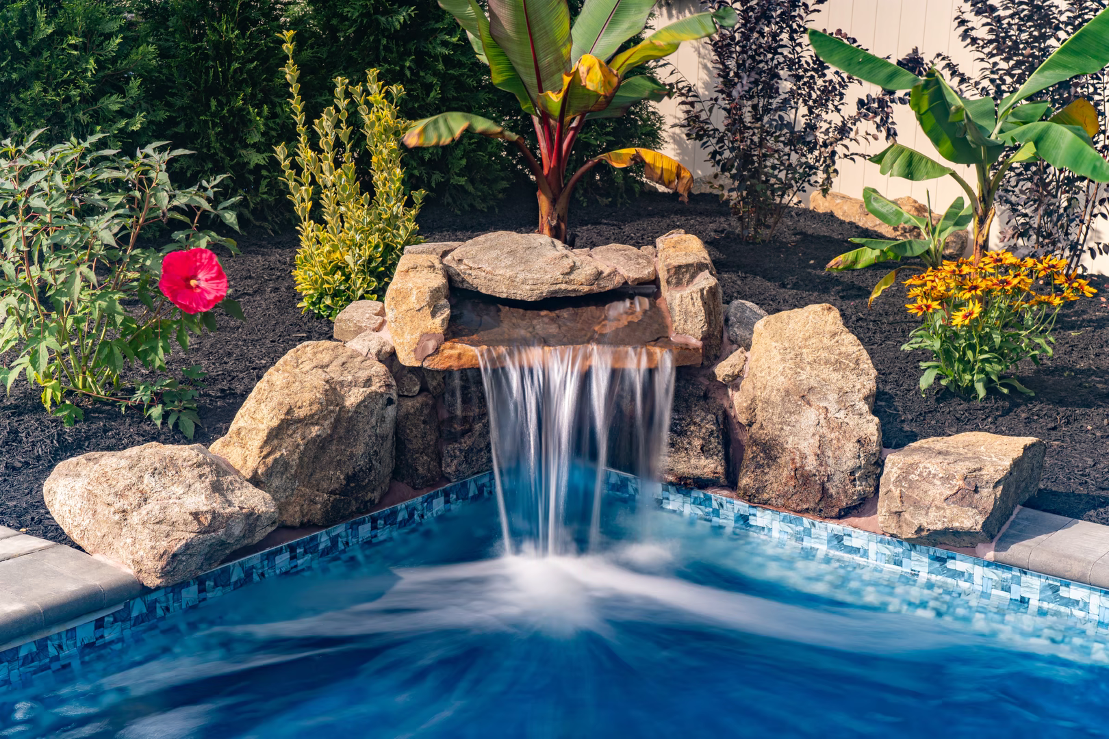 Genesis C - A small waterfall in the middle of a swimming pool surrounded by tropical plants and flowers