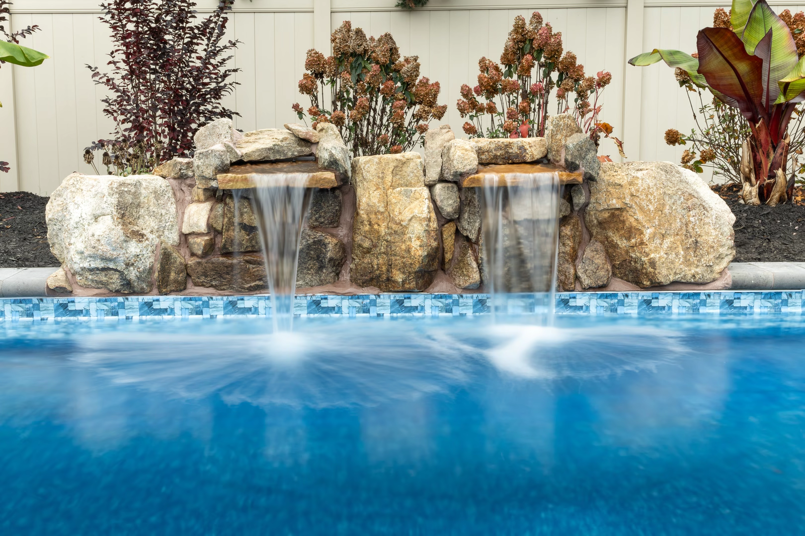 Dual natural stone boulder waterfalls flow into a tiled pool bordered by vibrant garden plants