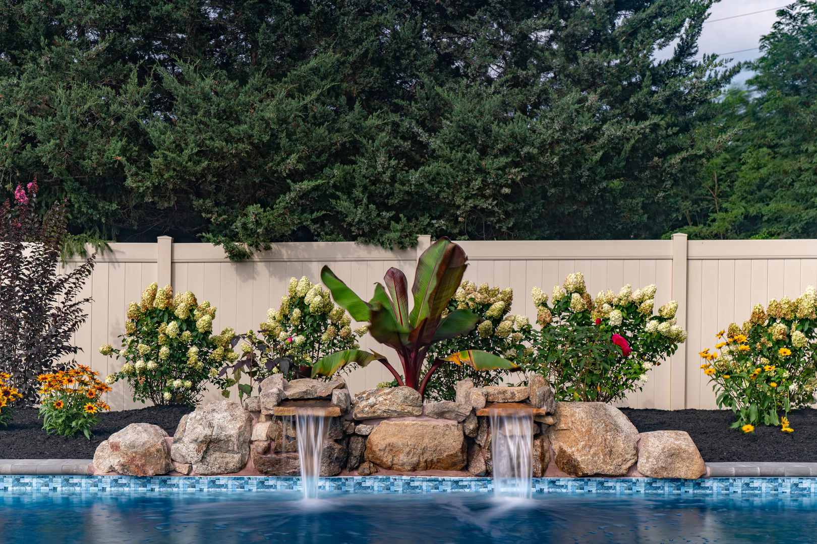 Twin natural stone boulder waterfalls cascading into a pool surrounded by lush garden plants