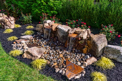 Genesis D - A garden with a small waterfall surrounded by rocks and plants.