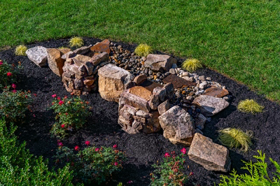 Natural stone boulder waterfall streambed feature in a mulched garden bed, framed by rose bushes, ornamental grasses, and lawn.