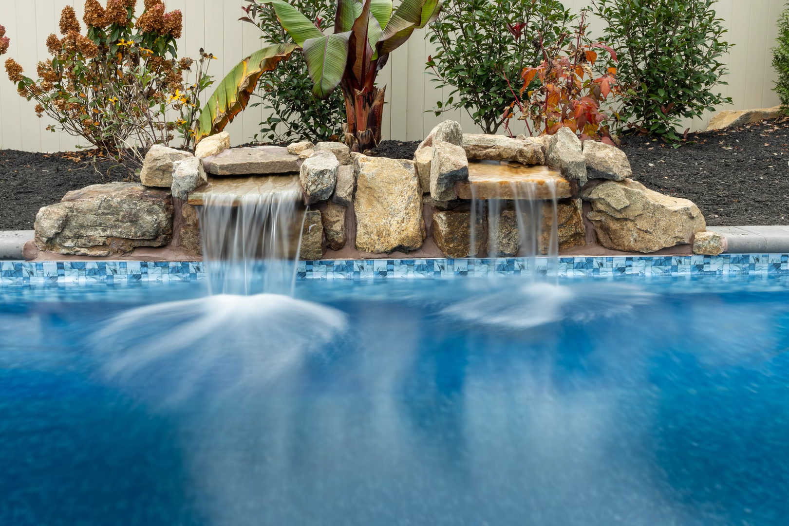 Twin natural stone boulder waterfalls cascading into a blue pool with lush garden plants