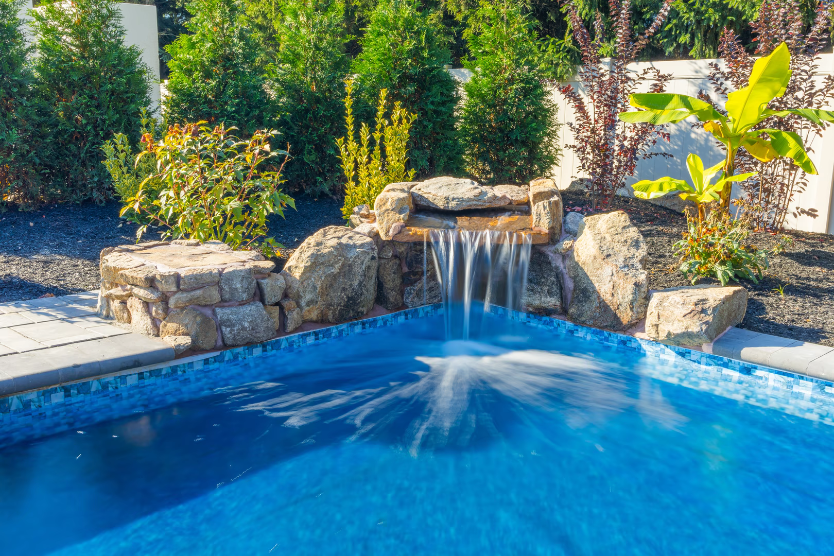 Genesis P - A swimming pool with a waterfall in the middle surrounded by lush greenery