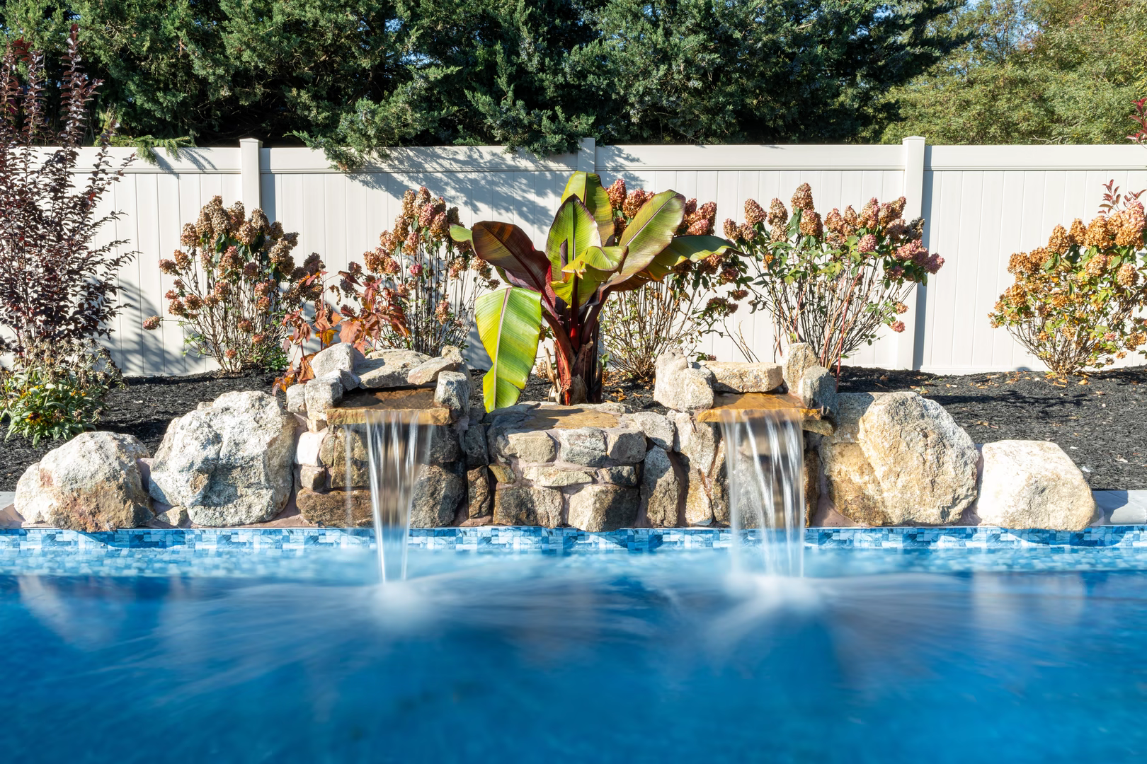Genesis P - A swimming pool with a waterfall feature surrounded by plants and a white fence
