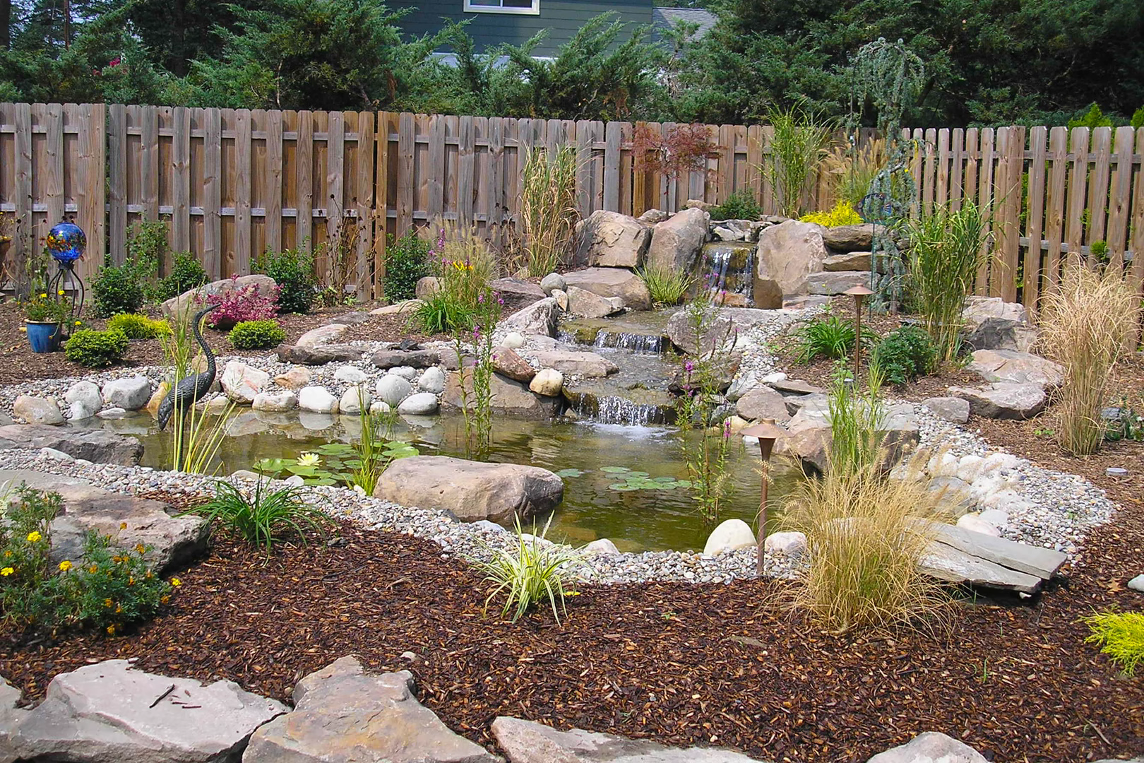 Natural stone boulder waterfall cascading into a pond surrounded by lush garden plants