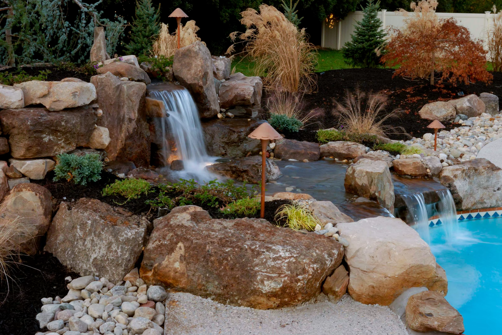 Two-tiered natural stone boulder waterfall cascading into a pool surrounded by ornamental plants