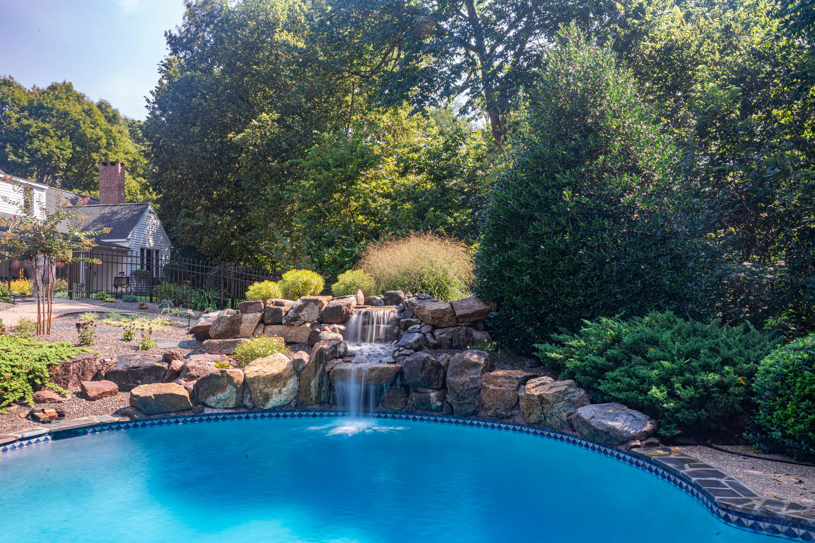 Natural stone boulder waterfall cascading into a backyard pool framed by lush greenery