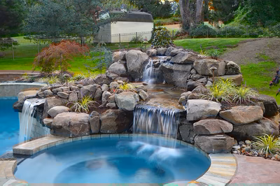 Himmilstein-F - A swimming pool lit up at night with a waterfall and rocks in the background