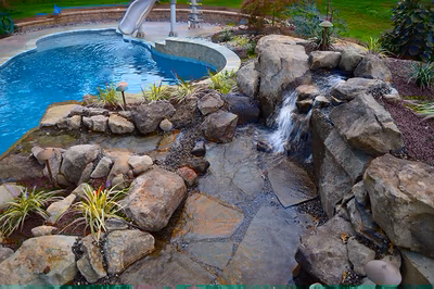 Himmilstein-G - A swimming pool lit up at night with a waterfall and rocks in the background