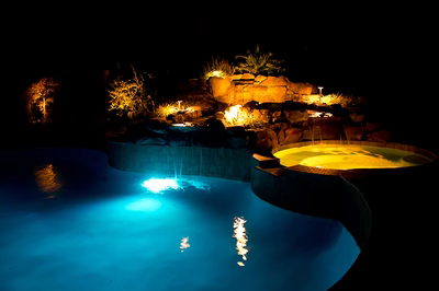 Himmilstein-K - A swimming pool lit up at night with a waterfall and rocks in the background