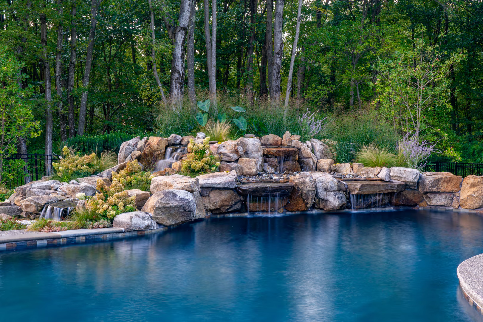 Natural stone boulder waterfalls cascading into a backyard pool surrounded by lush greenery
